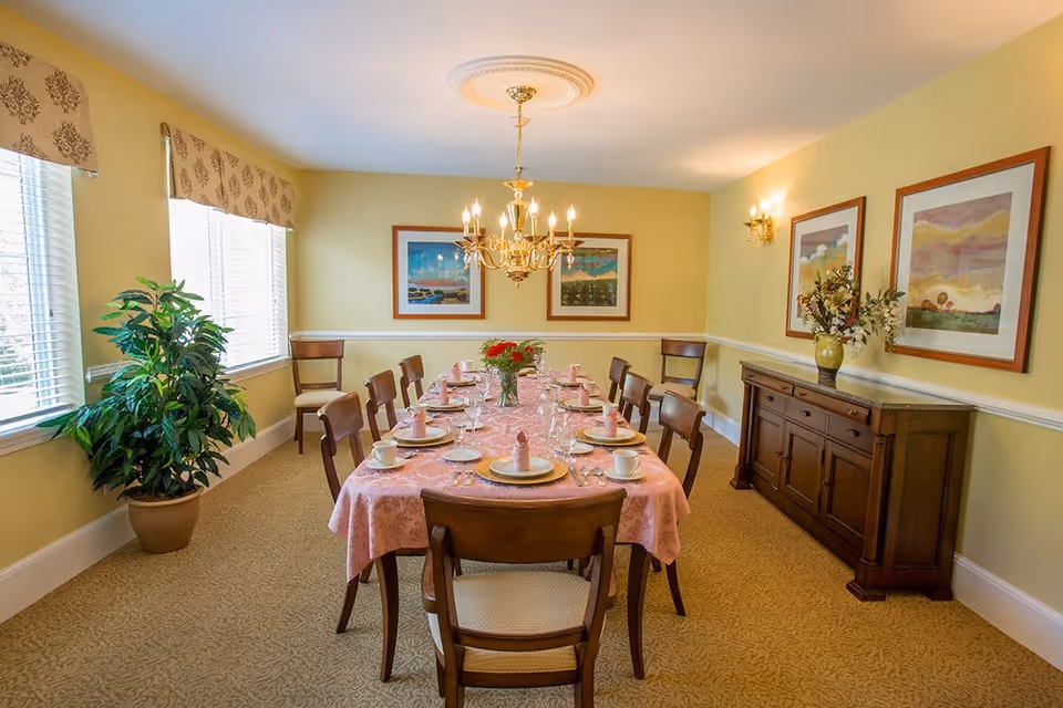 A dining room with a long table covered with a pink tablecloth set for a meal with plates, cups, napkins, and glasses. The room has yellow walls, a chandelier hanging from the ceiling, framed artwork on the walls, a wooden sideboard with a vase of flowers, and a large potted plant near two windows with blinds and valances.