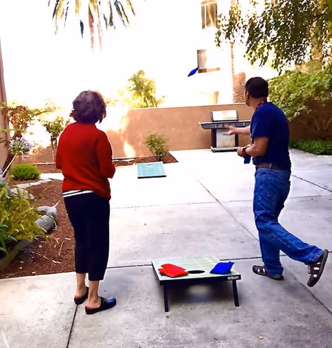 Two people playing cornhole outdoors on a concrete patio surrounded by plants and trees. One person is throwing a blue bean bag towards a cornhole board, while the other person watches. There is a barbecue grill in the background.
