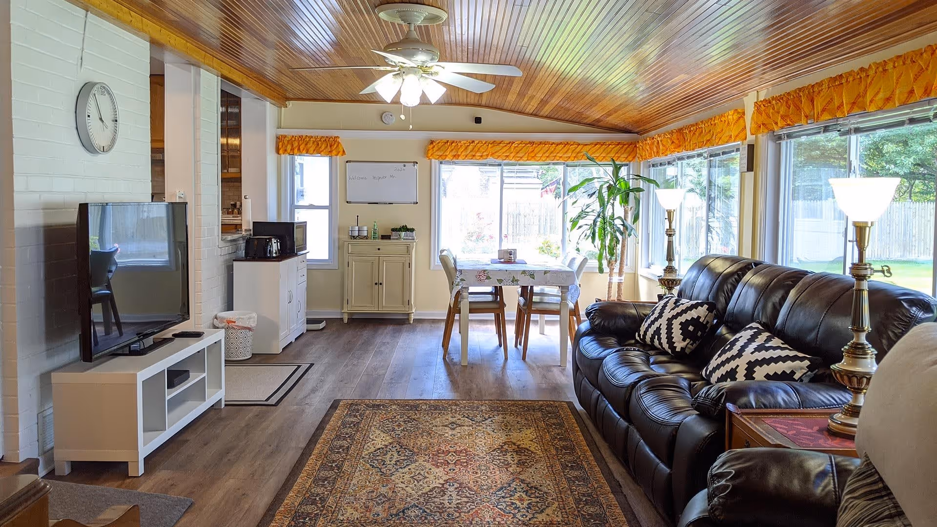 A bright living room with a wooden ceiling and large windows letting in natural light. The room features a black leather sofa with patterned cushions, two floor lamps, a patterned area rug, a TV on a white stand, and a small dining table with four chairs near the windows. There are plants and decorative items on cabinets along the walls.