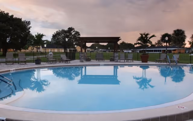 Outdoor swimming pool with clear blue water surrounded by lounge chairs and potted plants. In the background, there is a grassy area with trees, a wooden pergola, and a building under a cloudy sky at dusk.