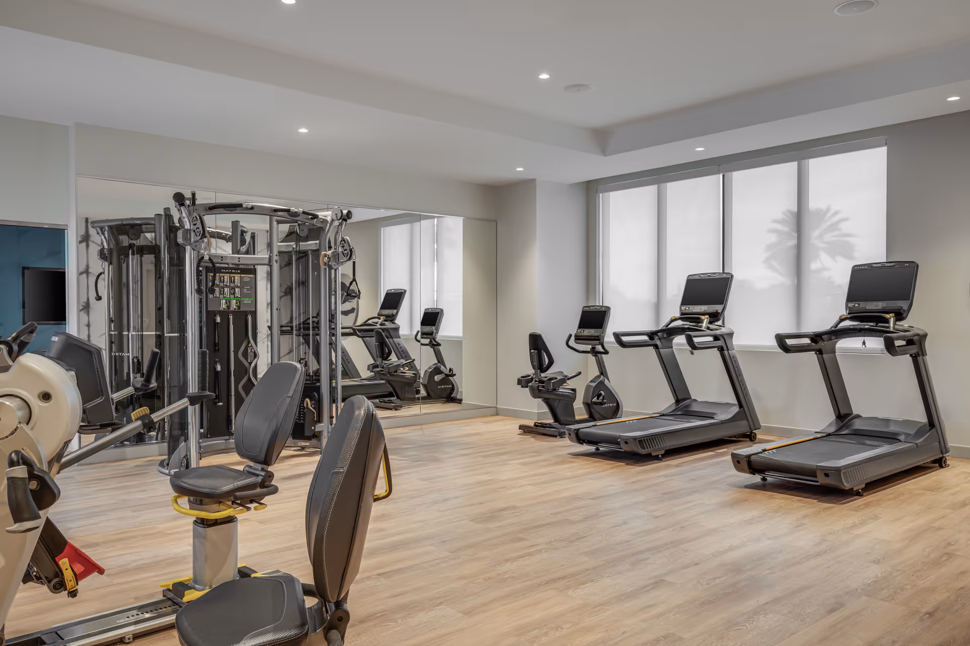 A modern fitness room with exercise equipment including treadmills, stationary bikes, and strength training machines. The room has wood flooring, large windows with blinds, and a large mirror on one wall.