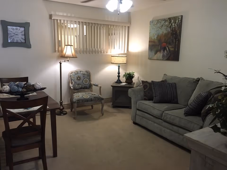 A cozy living room with a light green sofa adorned with striped and patterned pillows, a patterned armchair, a wooden side table with a lamp and a small plant, a floor lamp, a wooden dining table with chairs, a wall clock, vertical blinds on the window, and a painting of two people walking with umbrellas on a rainy path.