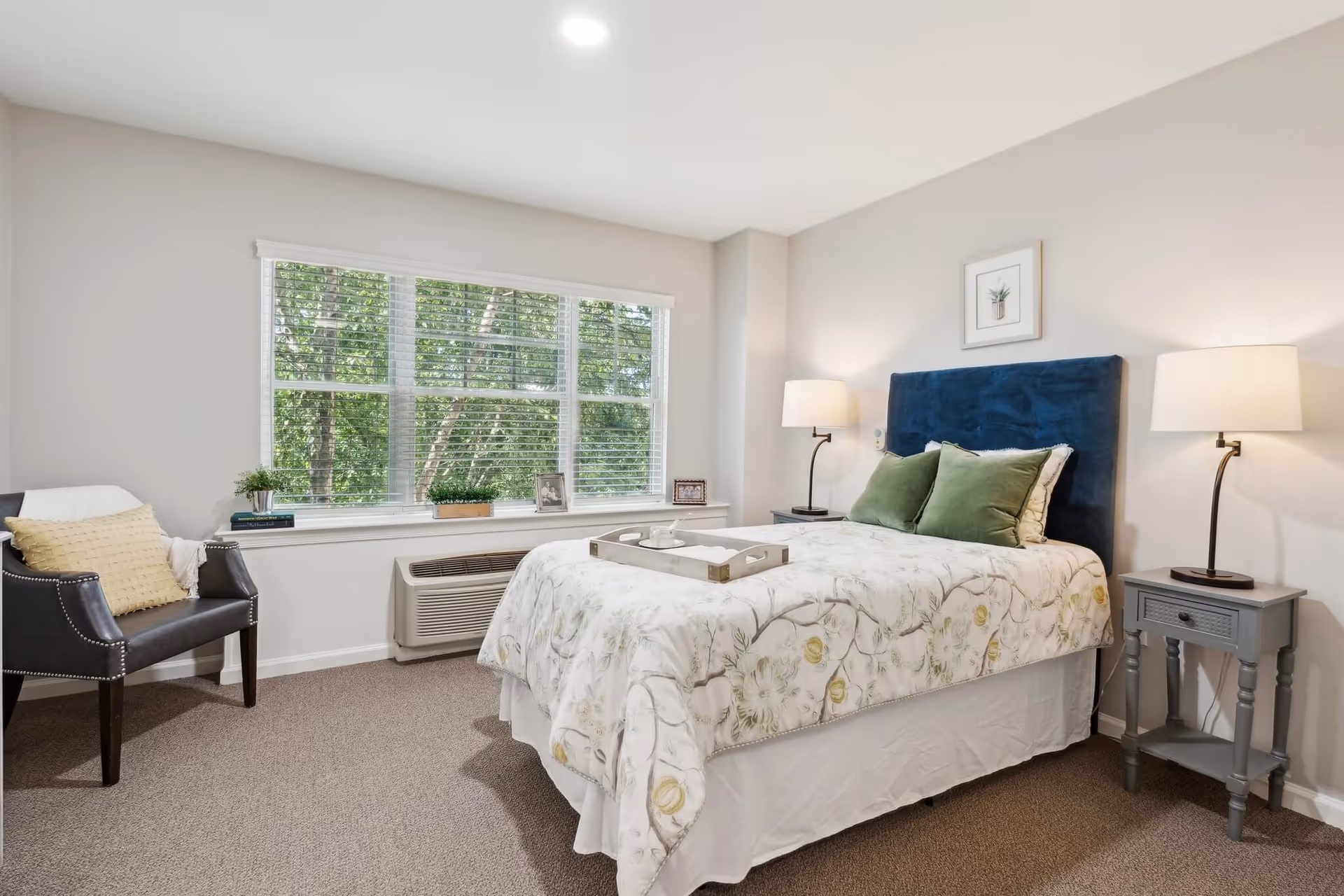 A bright and cozy bedroom featuring a bed with a floral-patterned comforter and two green pillows, flanked by two gray nightstands each with a lamp. There is a dark blue upholstered headboard and a framed picture above the bed. A window with white blinds lets in natural light, and a black armchair with a yellow pillow is placed near the window. The room has beige carpeting and light-colored walls.