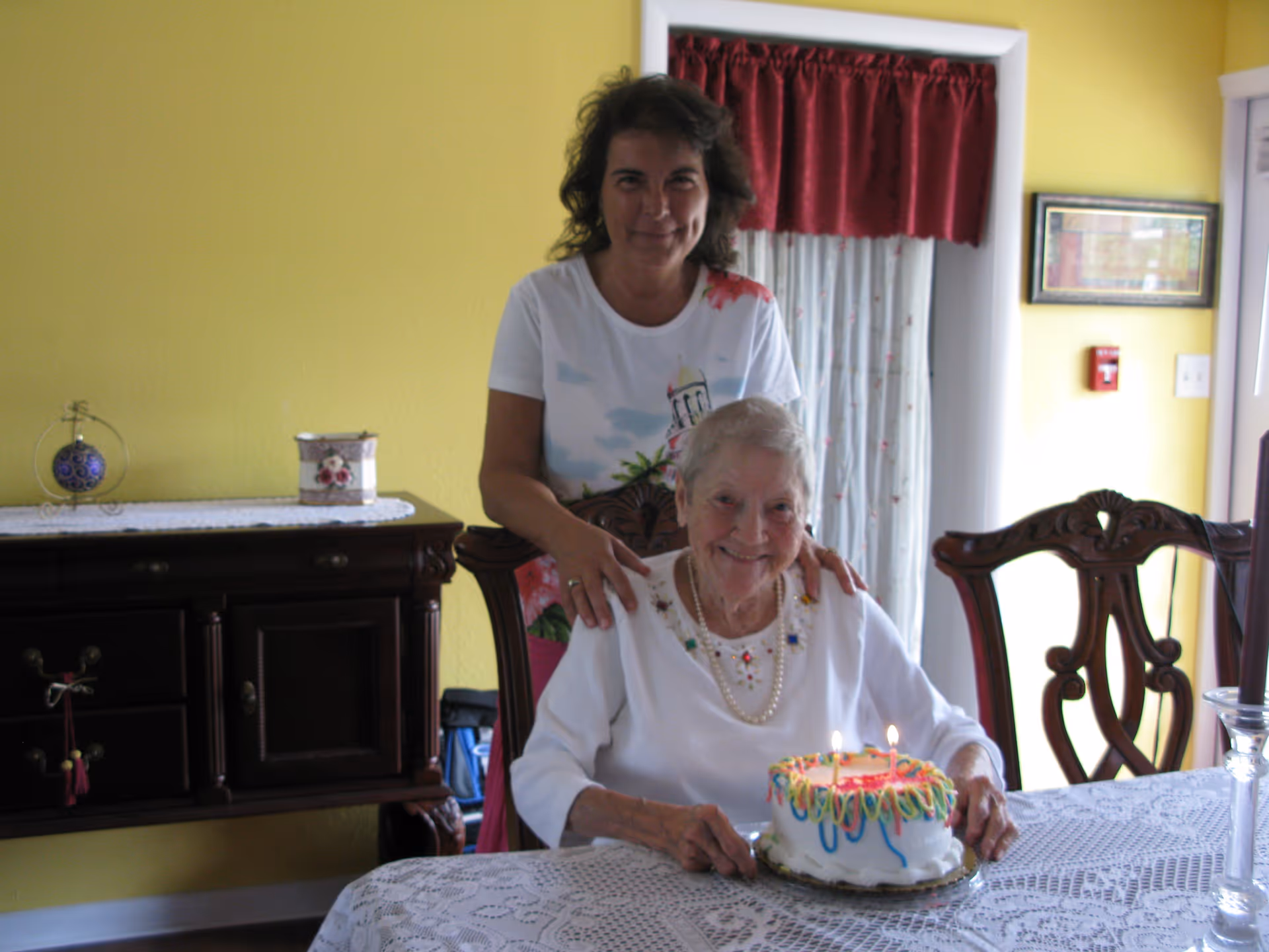 An elderly woman sitting at a dining table with a birthday cake with lit candles in front of her. A middle-aged woman stands behind her with hands on her shoulders. The room has yellow walls, a dark wooden sideboard, and a window with red curtains.