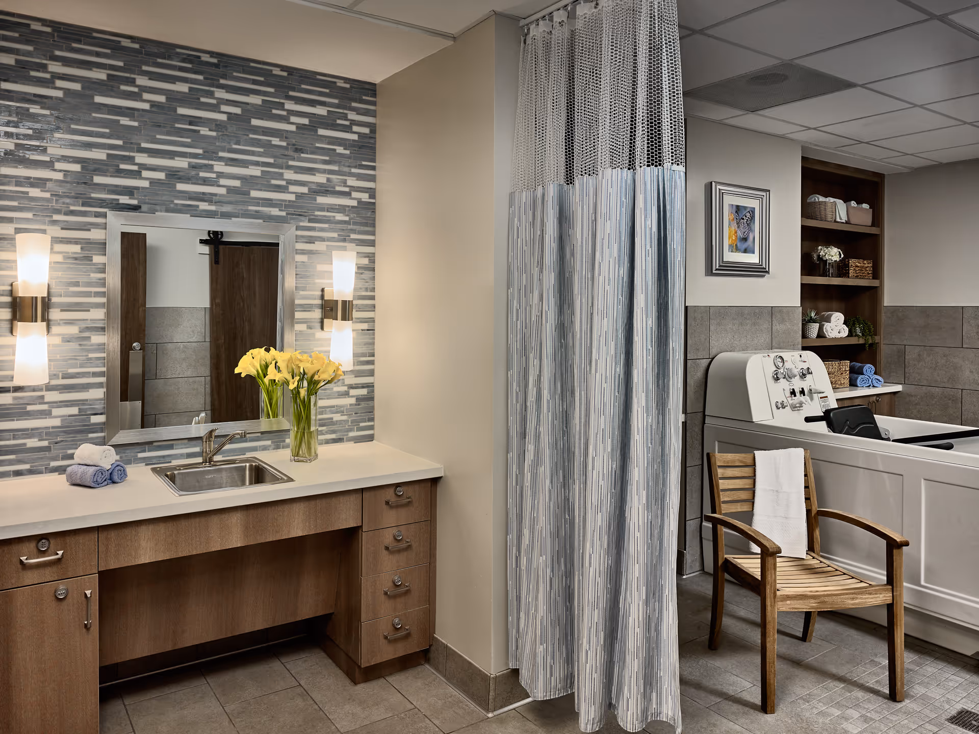 A clean and modern bathroom area with a wooden vanity featuring a sink, a mirror with two wall-mounted lights, and a vase of yellow flowers. To the right, there is a shower curtain and a wooden chair with a white towel draped over it. In the background, there is a large white bathtub with control knobs and a shelf with towels and decorative items.