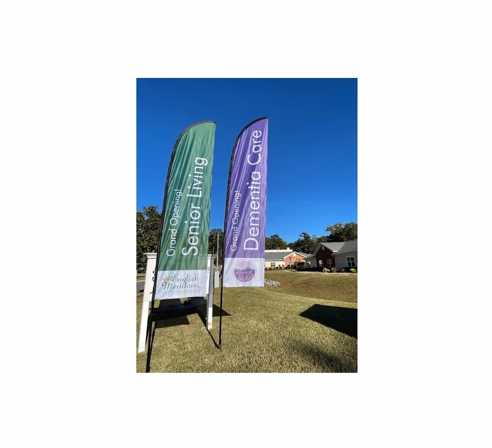 Two tall vertical flags on a grassy area under a clear blue sky. The green flag reads 'Grand Opening! Senior Living' with the English Meadows Senior Living Community logo at the bottom. The purple flag reads 'Grand Opening! Dementia Care'. In the background, there are buildings and trees.