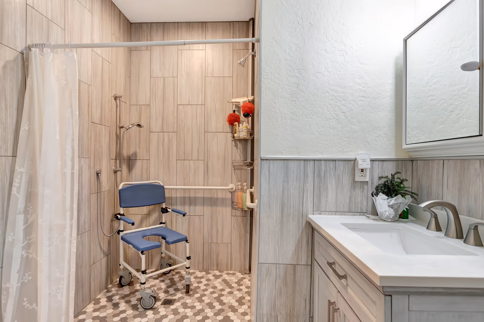 A bathroom with a walk-in shower featuring beige tiled walls and hexagonal floor tiles. Inside the shower is a blue and white shower chair with wheels and grab bars for accessibility. There is a shower curtain on the left side and a wall-mounted showerhead. To the right is a white sink vanity with a silver faucet, a small potted plant, and a mirrored medicine cabinet above.