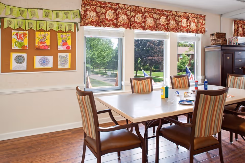 A bright room with a large table surrounded by six striped cushioned chairs. On the table are art supplies including paint bottles and a paint palette. The room has three large windows with a view of green trees and a street outside. A bulletin board on the wall displays colorful artwork and a banner that reads 'LIVE WITH PURPOSE'.