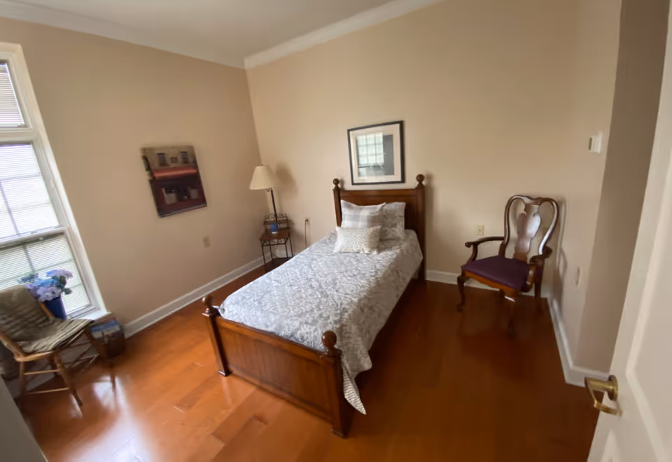 A small bedroom with a single wooden bed covered with a patterned white and gray bedspread and two pillows. There is a wooden chair with a purple cushion to the right of the bed, a floor lamp and small table with a blue item on it to the left, and a wicker chair with a cushion near a window with blinds. The walls are beige, and there is a framed picture above the bed and another picture on the left wall. The floor is wooden.