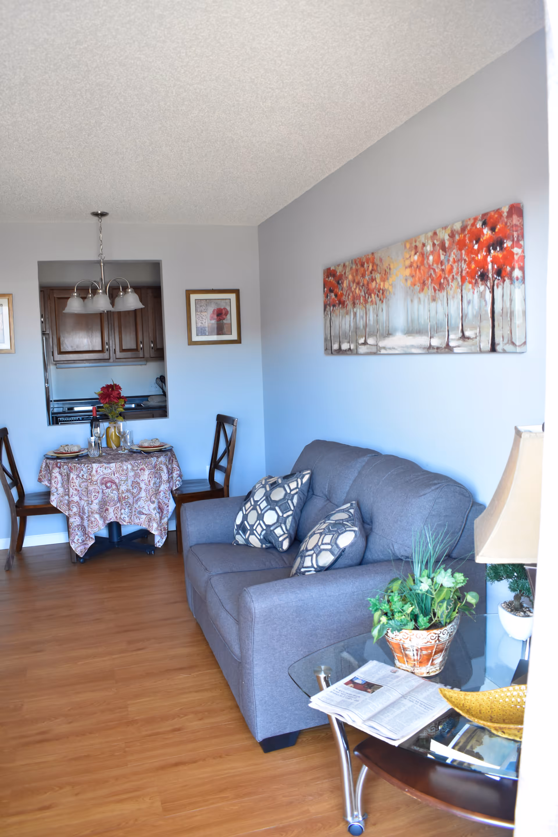 A cozy living room area with a gray loveseat adorned with two patterned pillows. Next to the loveseat is a glass side table holding a potted plant, a lamp, a newspaper, and a decorative bowl. On the wall above the loveseat is a painting of trees with red and orange leaves. In the background, there is a small dining area with a round table covered with a patterned tablecloth, set for two with plates, glasses, and a vase with red flowers. The dining area is adjacent to a kitchen with wooden cabinets and a pass-through window.