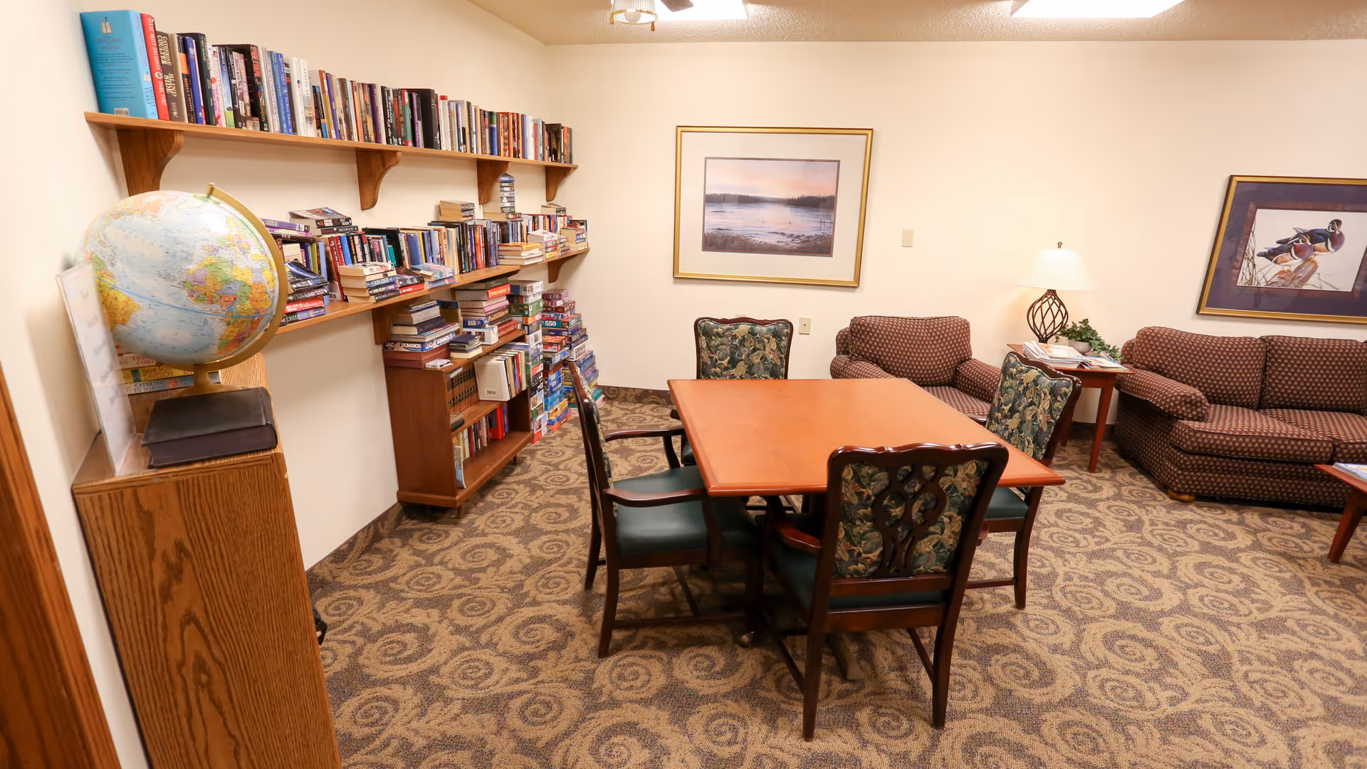 A cozy room with a wooden table surrounded by four chairs with floral upholstery. There are two patterned sofas, a side table with a lamp, and framed artwork on the walls. A bookshelf filled with books and board games lines one wall, and a globe sits on a wooden stand near the bookshelf. The carpet has a swirling pattern.