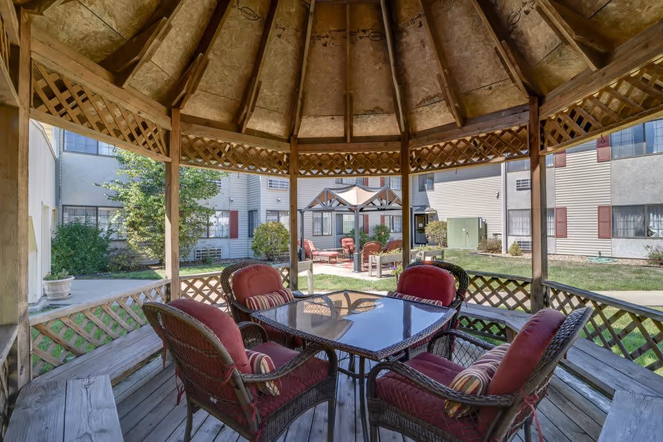 View from inside a wooden gazebo with a glass-top table and four cushioned wicker chairs with red cushions and striped pillows. The gazebo overlooks a courtyard with grass, shrubs, and another seating area with red chairs and a canopy. Surrounding the courtyard are multi-story residential buildings with beige siding and red shutters.