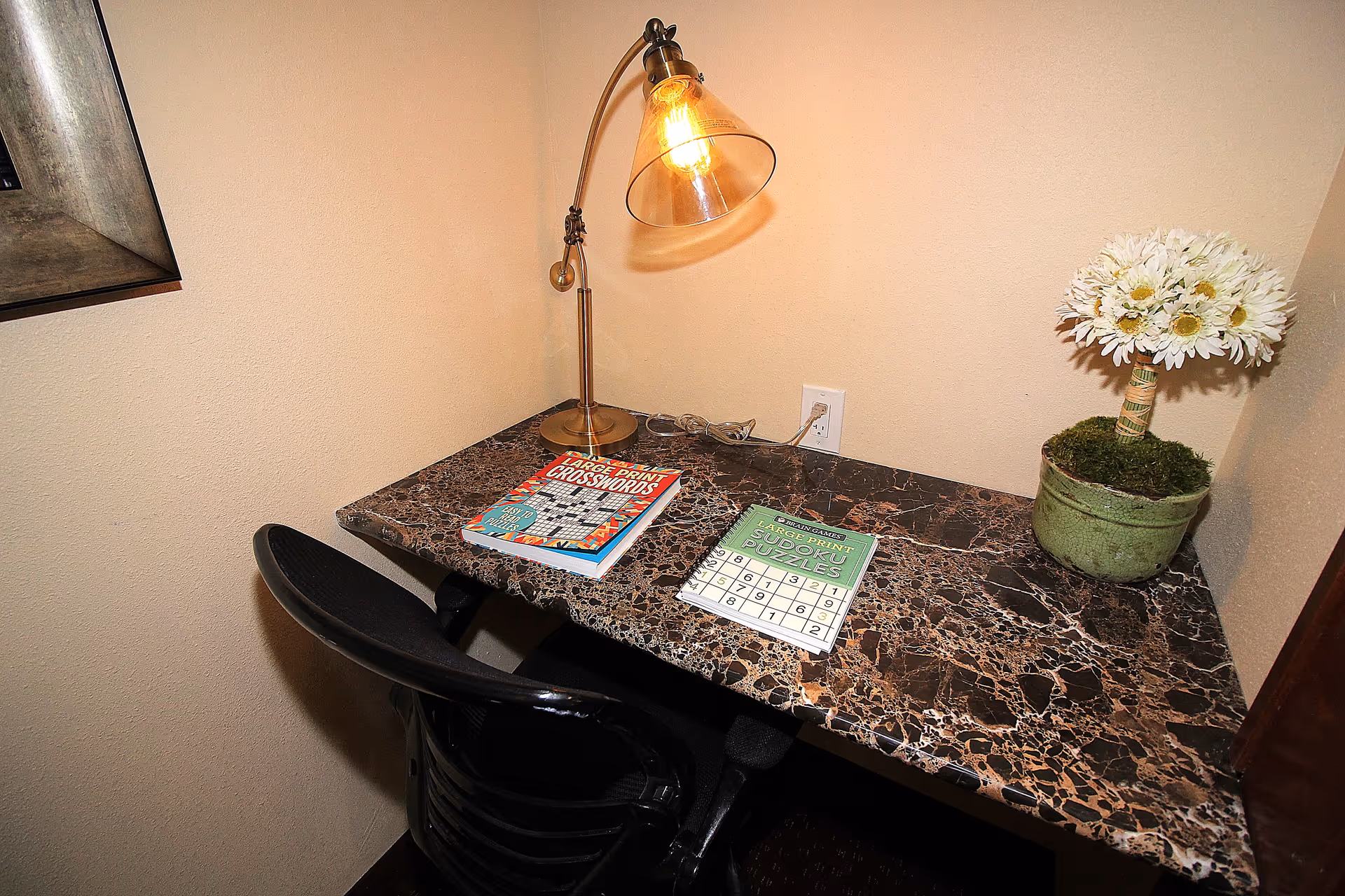 A small marble-patterned desk with a black chair, a brass desk lamp with a clear glass shade, two puzzle books titled 'Large Print Crosswords' and 'Large Print Sudoku Puzzles', and a small potted plant with white flowers on the right side of the desk.