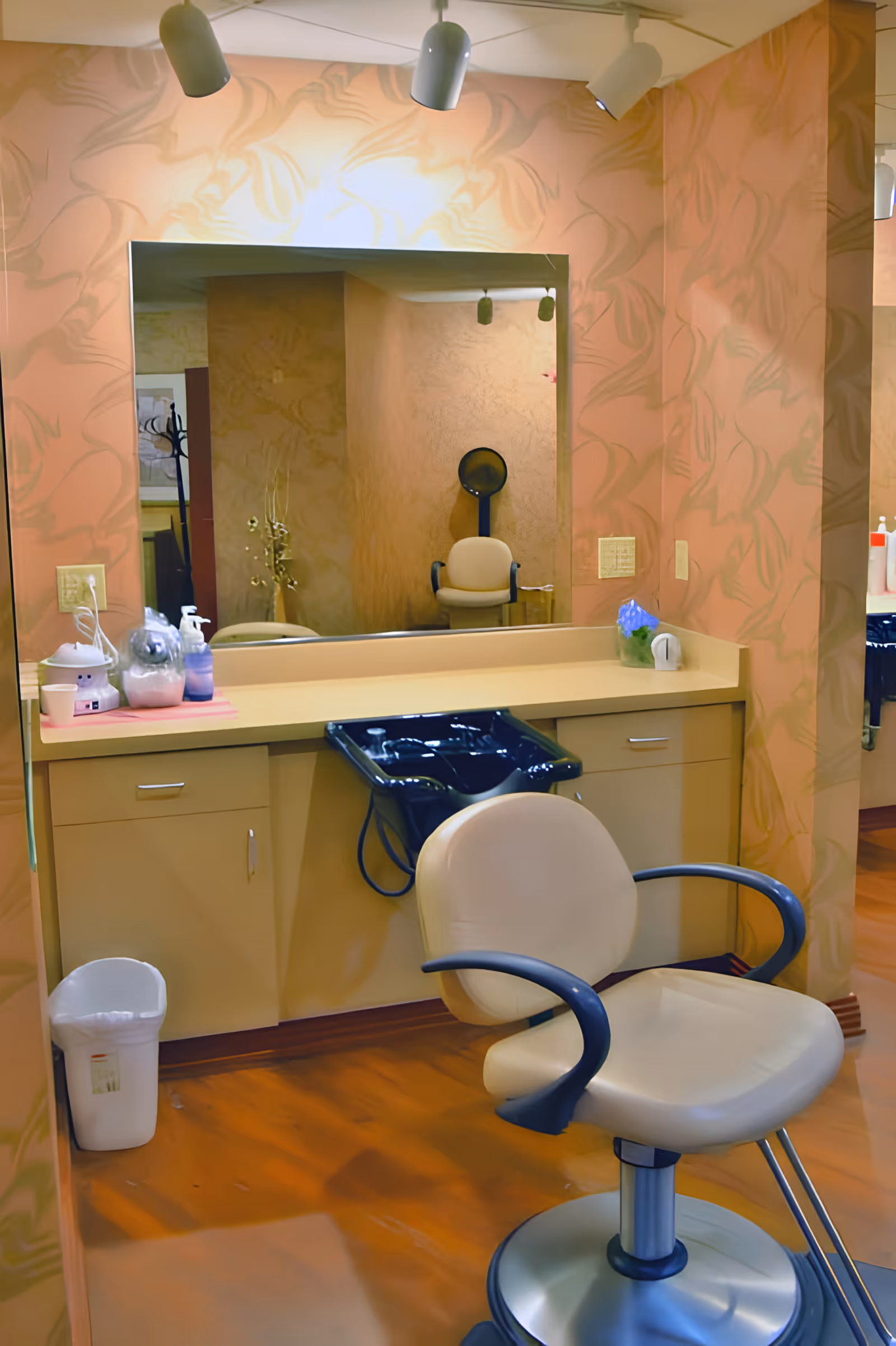 Interior view of a hair salon station with a beige salon chair in front of a counter with a black hair washing sink. The wall has a large mirror and is decorated with a subtle floral pattern. Various hair care products and a trash bin are visible.