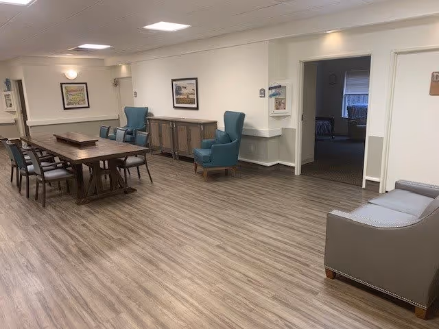 A spacious interior room with wood flooring featuring a long wooden dining table surrounded by chairs, two teal armchairs, a gray sofa, and a wooden sideboard against the wall. The walls are light-colored with framed artwork and there is an open doorway leading to another room.