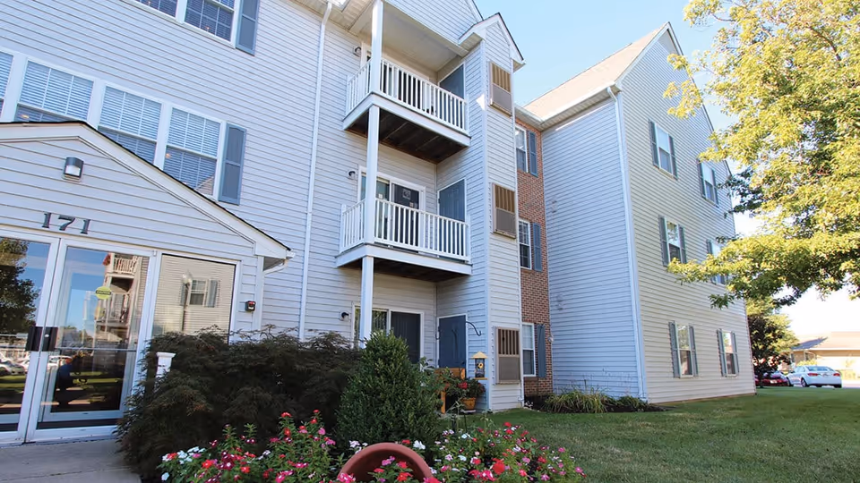Exterior view of a three-story residential building with balconies, an entrance and landscaped front lawn with flowers.