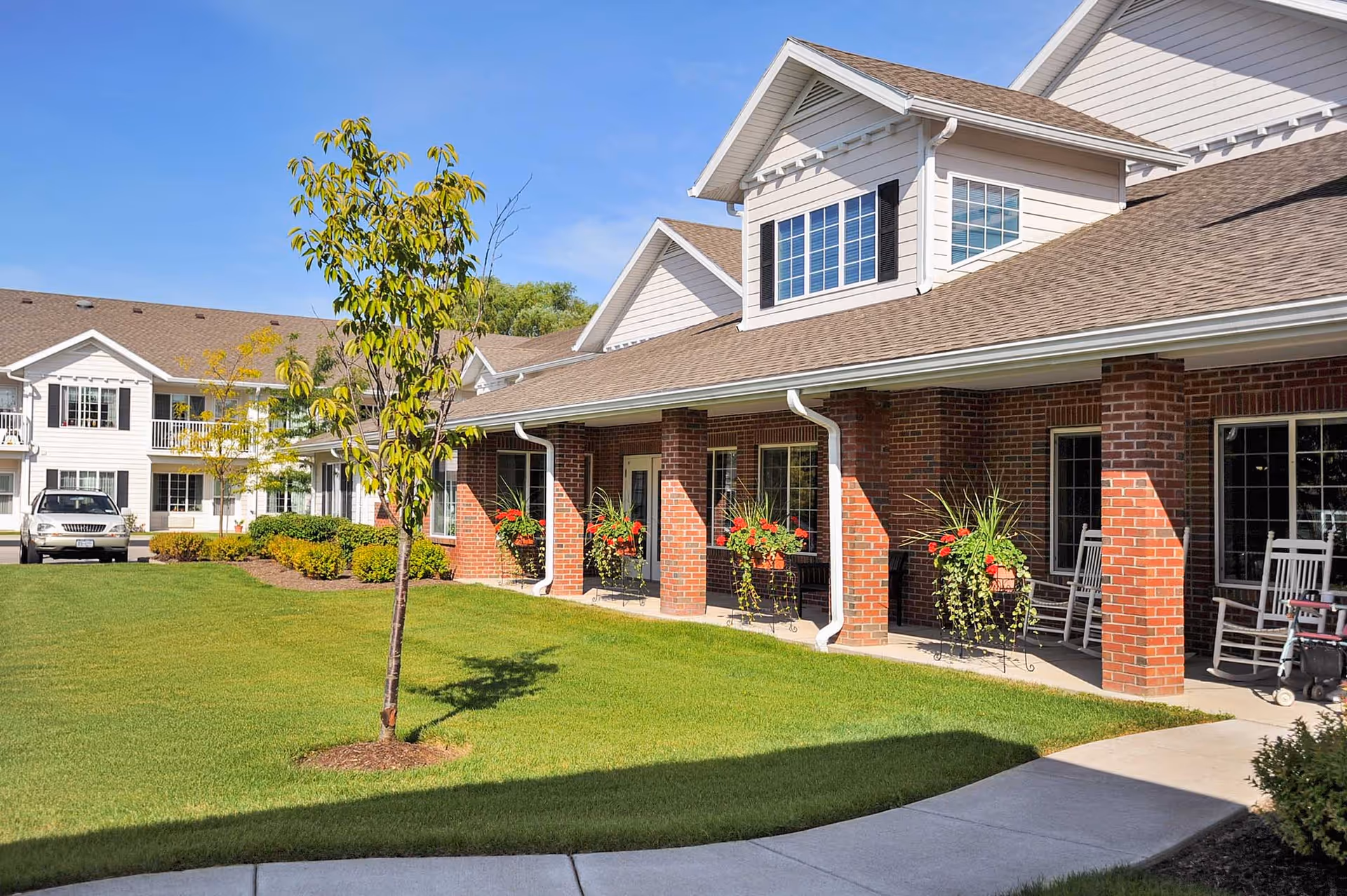 Exterior view of Holiday Fleming Point facility showing a well-maintained lawn, a young tree, and a building with brick pillars and white siding. The building has multiple windows, flower pots with red flowers, and rocking chairs on the covered porch. A silver SUV is parked near the building under a clear blue sky.