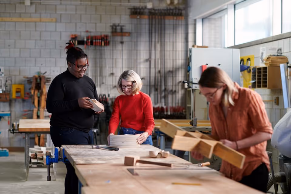 Three people working on woodworking projects at a long workbench in a bright workshop.