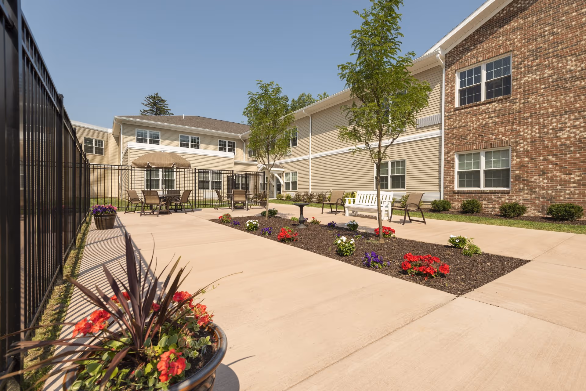 Sunlit courtyard with patio seating, planters, and a landscaped walkway beside a two-story assisted living building.