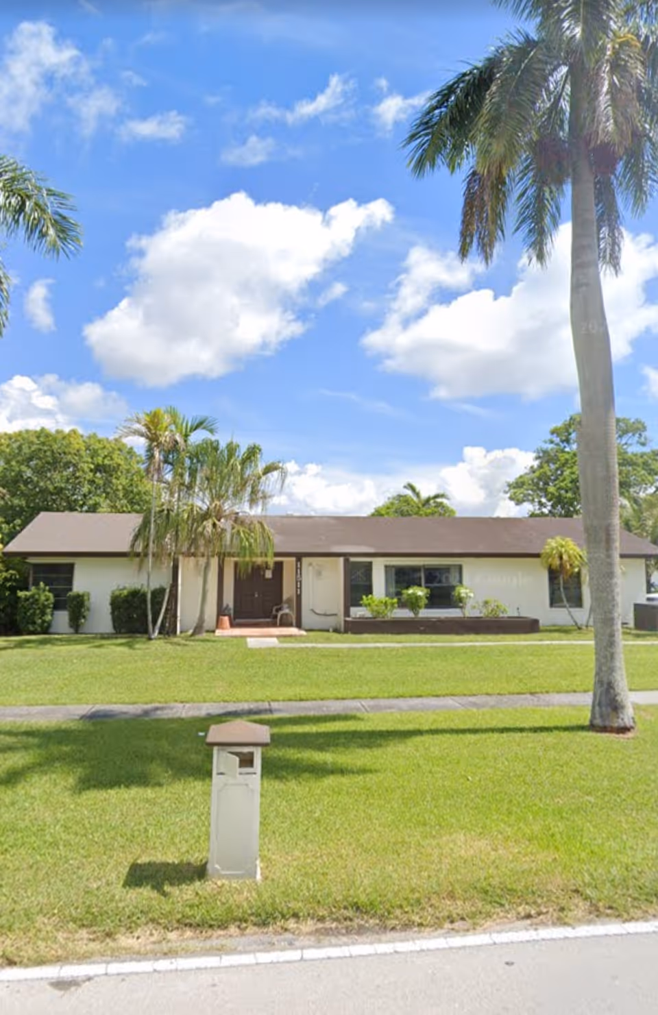 Single-story building front with a manicured lawn, palm trees, and a mailbox under a blue sky.