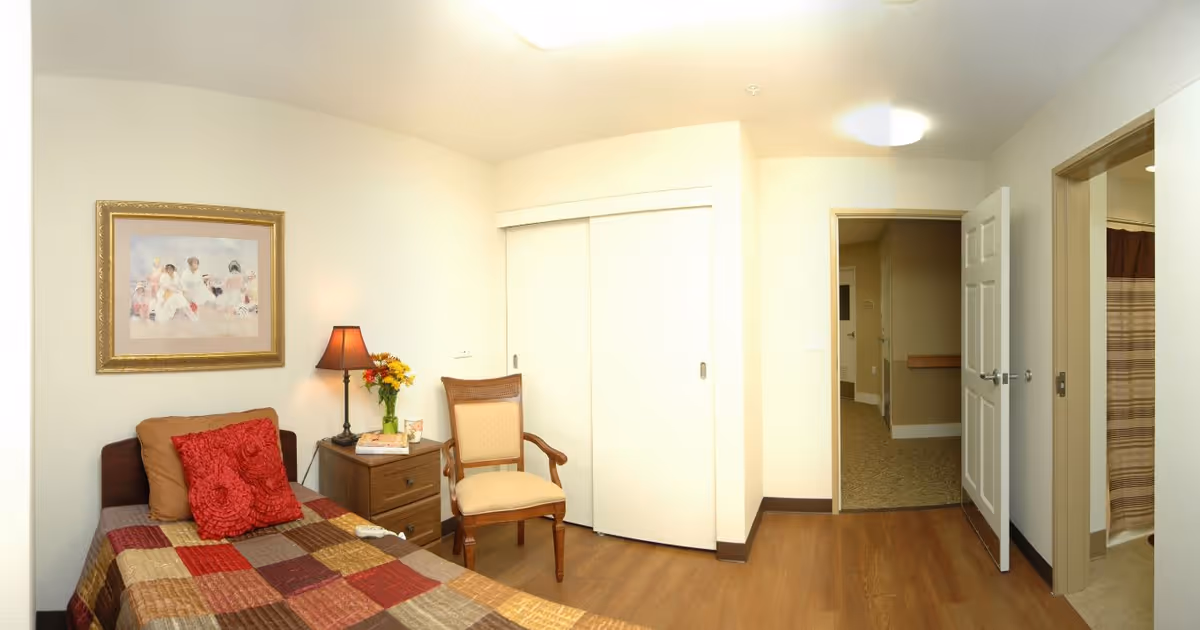 A small bedroom with a single bed covered in a patchwork quilt with red and brown tones. There are two red decorative pillows and one brown pillow on the bed. Next to the bed is a wooden nightstand with a lamp, a vase of flowers, and some books. A wooden chair with a cushioned seat is placed beside the nightstand. The room has light-colored walls, a framed painting above the bed, a closet with sliding white doors, and an open door leading to a hallway and a bathroom with a striped shower curtain.