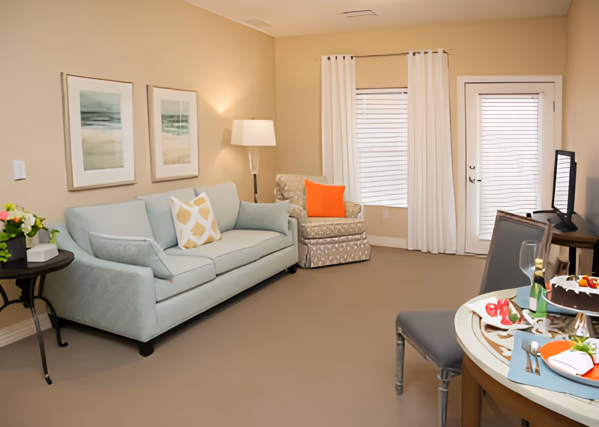 A cozy living room with a light blue sofa adorned with a patterned pillow, a floral armchair with an orange pillow, a floor lamp, two framed abstract paintings on the wall, and a round table set with plates, glasses, and a cake. The room has beige walls, a window with white curtains, and a door with blinds.