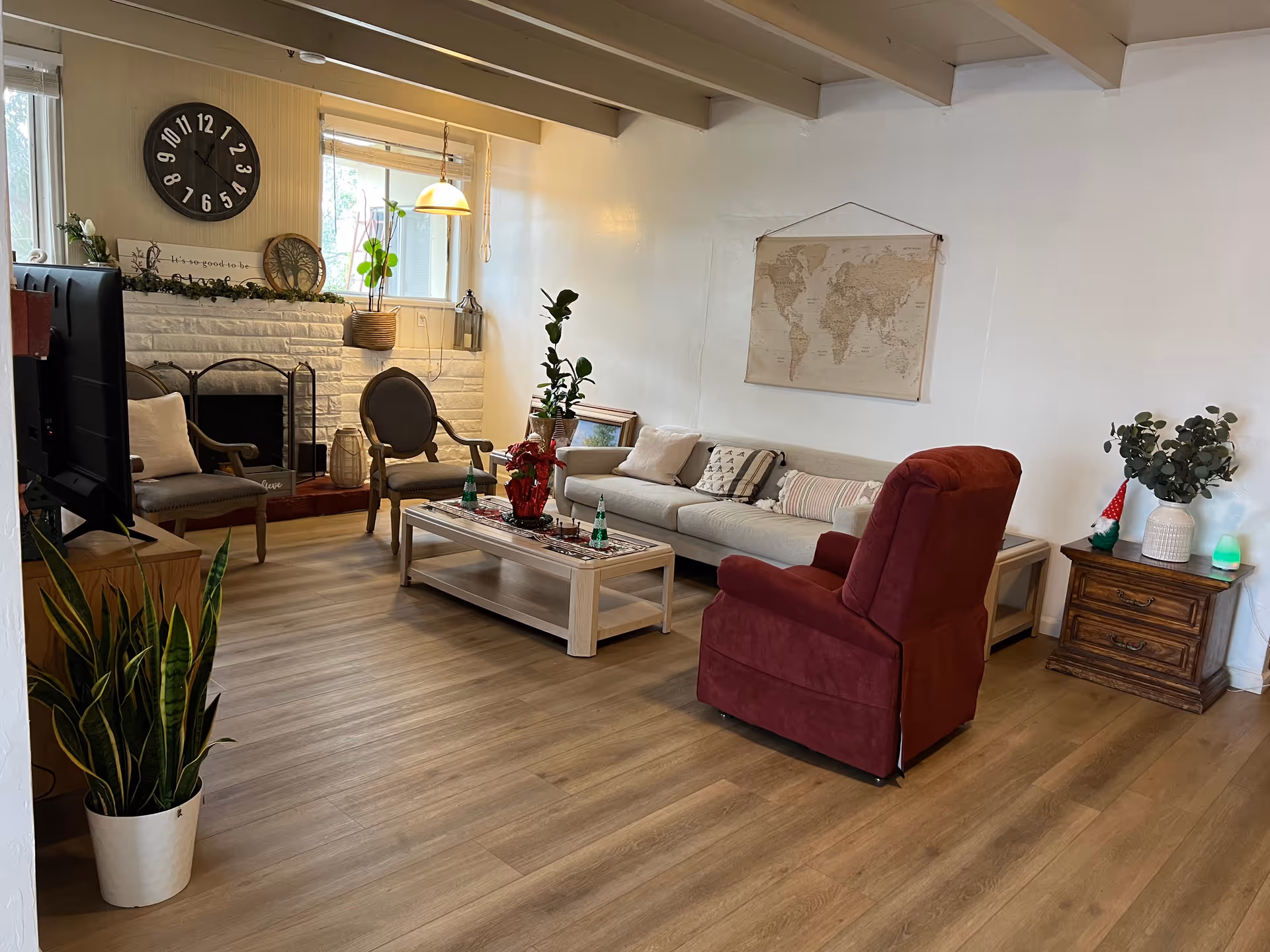 A cozy living room with a beige sofa, a red armchair, two wooden chairs with cushions, a white coffee table decorated with plants and small ornaments, a large wall clock above a white brick fireplace, a world map hanging on the wall, and several potted plants placed around the room. The floor is wooden and the room is well-lit by natural light from a window.