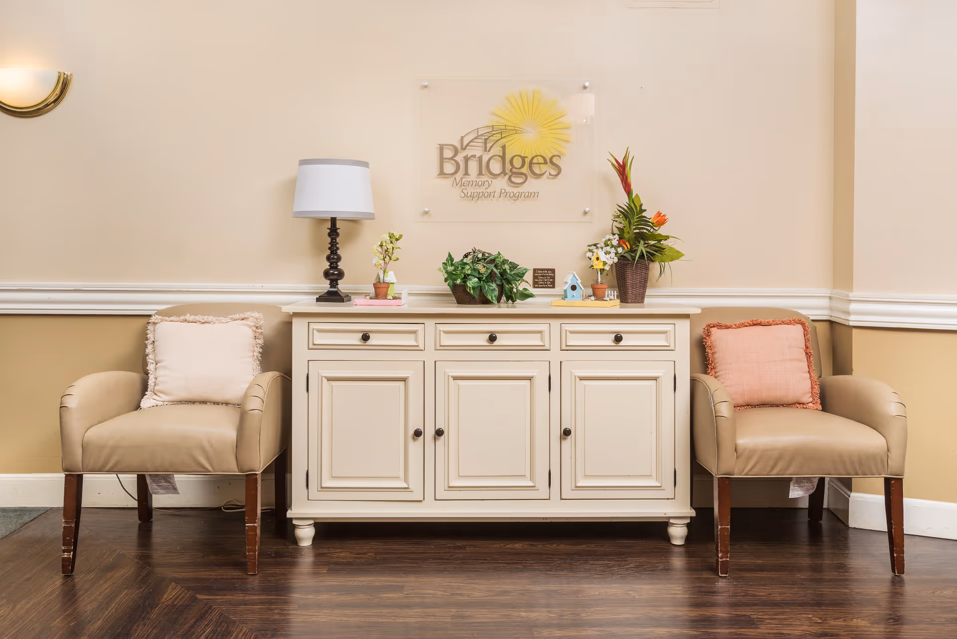 A cozy seating area with two beige armchairs flanking a cream-colored cabinet topped with a lamp, plants and a 'Bridges Memory Support Program' sign.