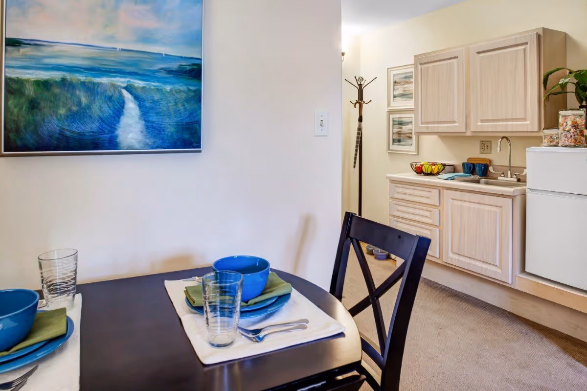 Small dining area with a set table and blue dishes next to a kitchenette with light wood cabinets and a mini-fridge.