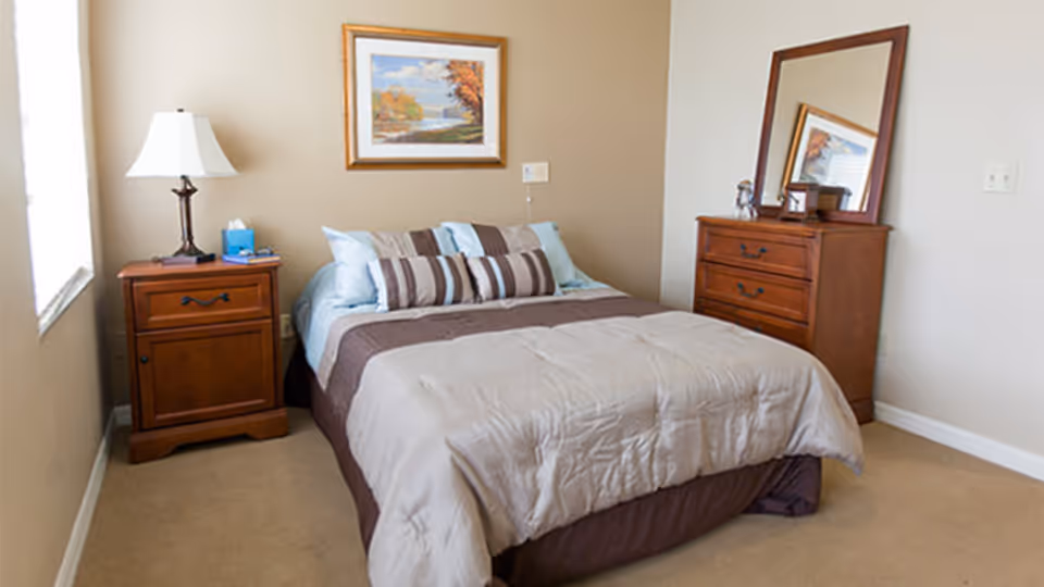 A neatly made bedroom with a double bed, wooden nightstand and dresser, lamp, and framed artwork on the wall.