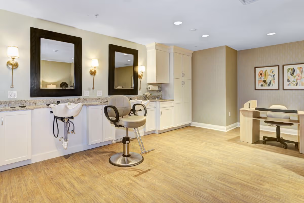 Bright interior salon area with two mirrors, a styling chair and sink, white cabinetry, and a small desk against a wood floor.