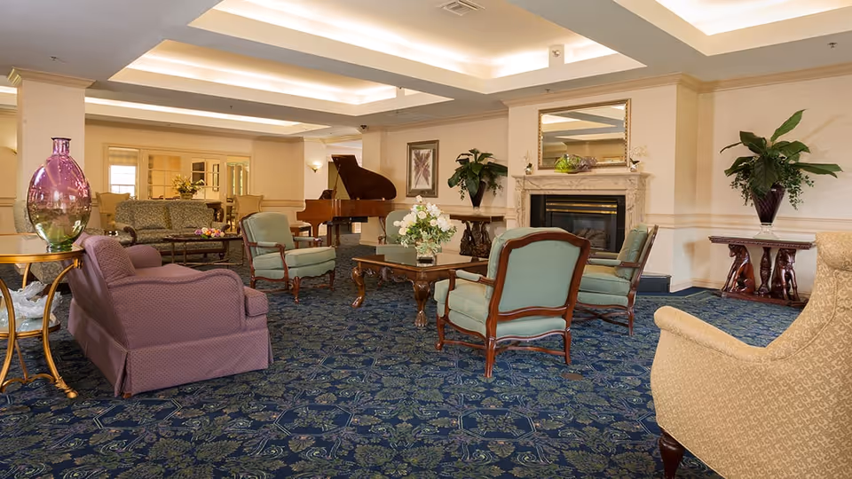 A spacious and elegantly furnished living room area with patterned blue carpet, several upholstered chairs and sofas arranged around a wooden coffee table with a floral centerpiece. There is a grand piano in the background, a fireplace with a mirror above it, and decorative plants and artwork on the walls.