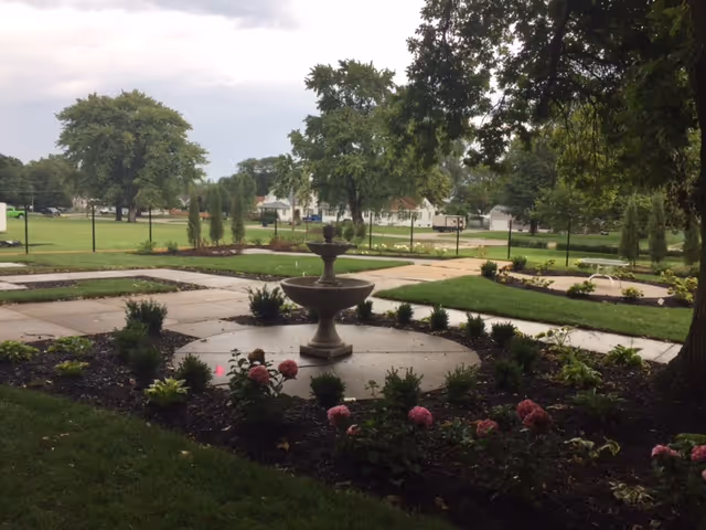 Landscaped courtyard with a central tiered fountain, flower beds, paved walkways, and surrounding trees and lawn.