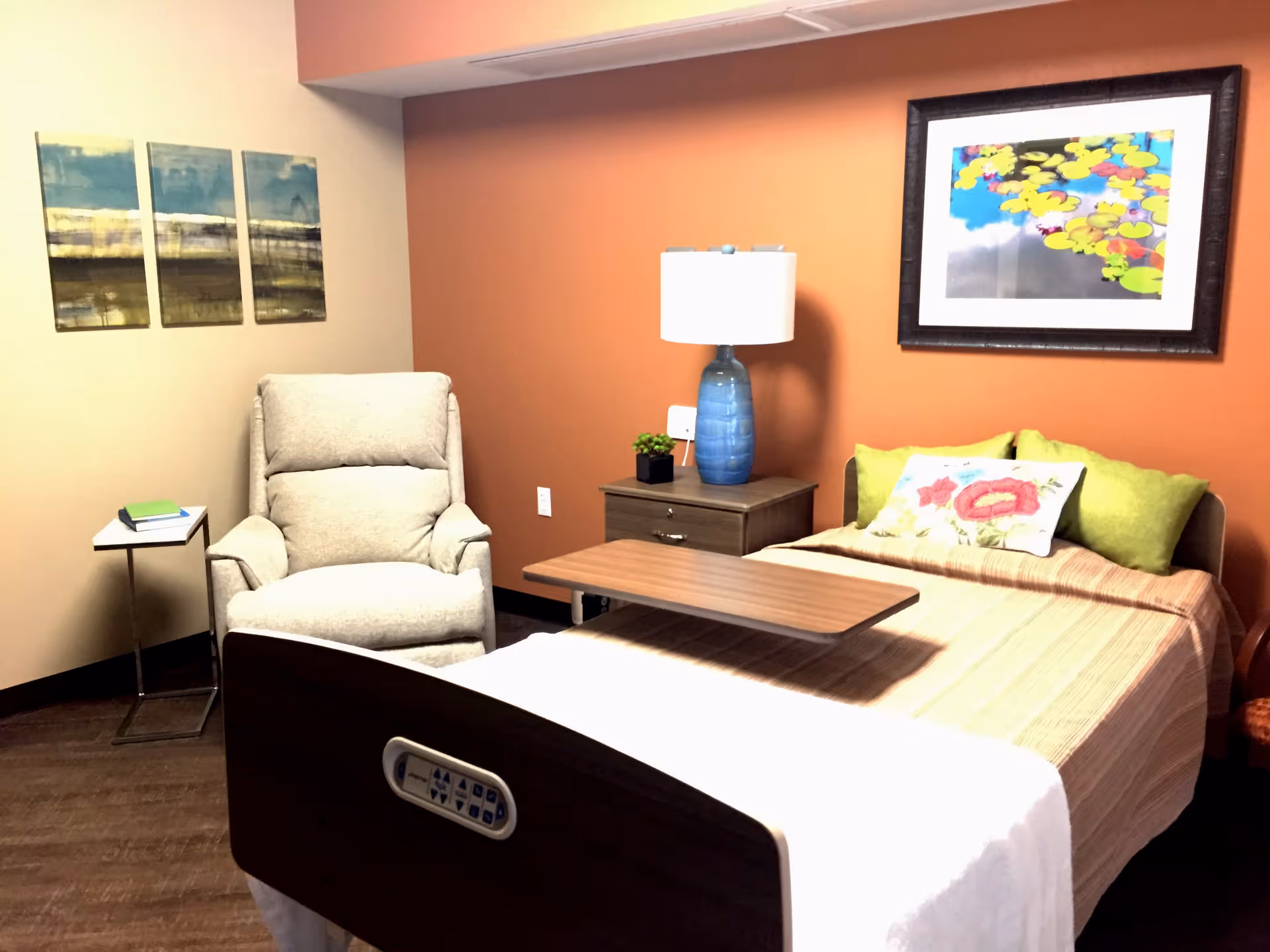 A cozy rehabilitation suite featuring a hospital-style bed with a wooden overbed table, a beige recliner chair, a small side table with books, a nightstand with a blue lamp and a small plant, and two pieces of wall art. The walls are painted in warm tones with one wall in orange and the others in beige.