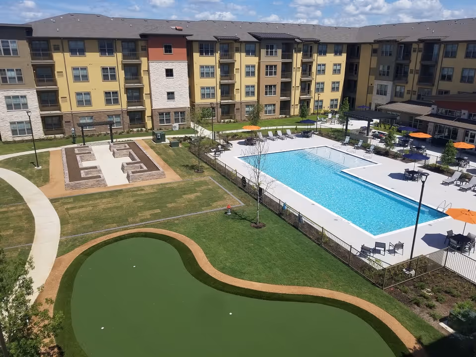 Outdoor view of a senior living facility courtyard featuring a swimming pool with lounge chairs and umbrellas, a putting green, and a fire pit area surrounded by a multi-story residential building under a partly cloudy sky.