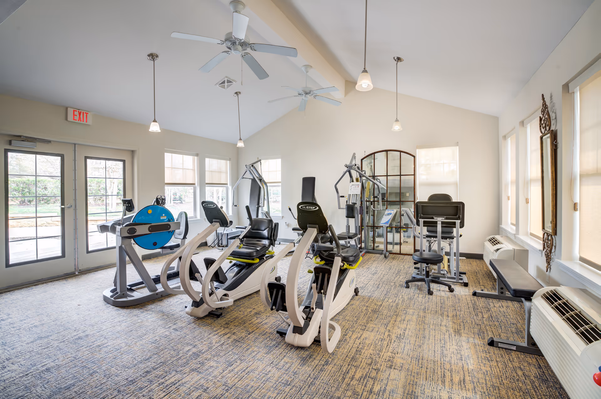 Bright fitness room with exercise equipment including stationary bikes, weight machines, and a bench. The room has large windows with shades, ceiling fans, pendant lights, and a door leading outside.