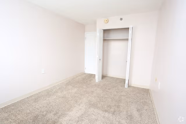 Empty room with beige carpet, light-colored walls, an open closet with white doors, and a closed white door.