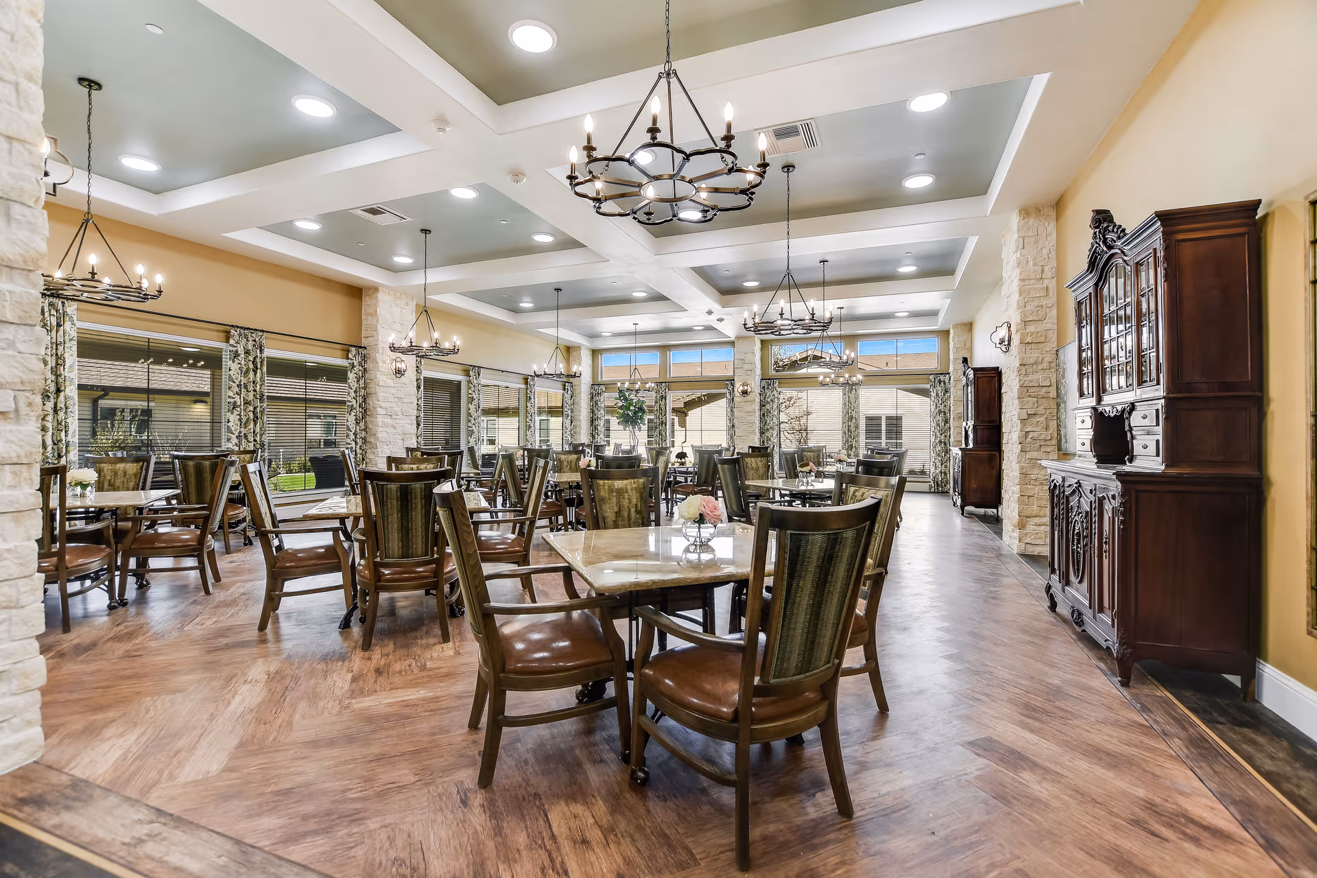 A spacious senior living dining room with multiple wooden tables and chairs arranged neatly. The room features large windows with floral curtains, stone pillars, and elegant chandeliers hanging from a coffered ceiling. The floor has a wood pattern, and there are dark wooden cabinets along the walls.