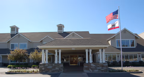 Front exterior view of a senior living facility building with a covered entrance supported by white columns and stone bases. Two flagpoles stand in front, flying the American flag and the California state flag. The building has multiple windows and a sloped roof with dormer windows.