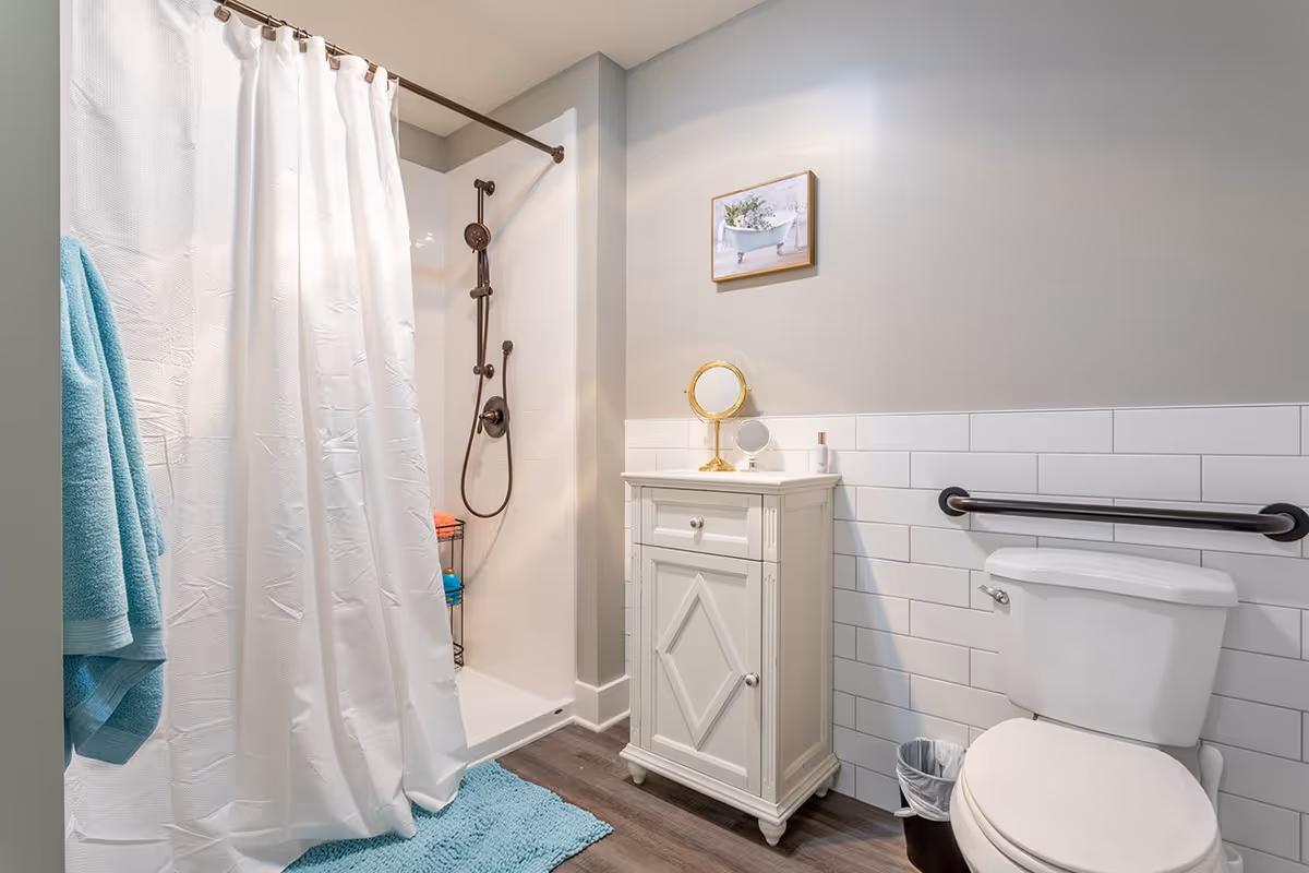 A clean and modern bathroom featuring a white shower with a bronze showerhead and a white curtain, a white cabinet with a small round mirror on top, a white toilet with a black grab bar mounted on the wall beside it, a small trash can, and a blue towel hanging on a hook. The walls are painted light gray with white subway tile halfway up, and there is a framed picture of a bathtub with flowers hanging above the cabinet.