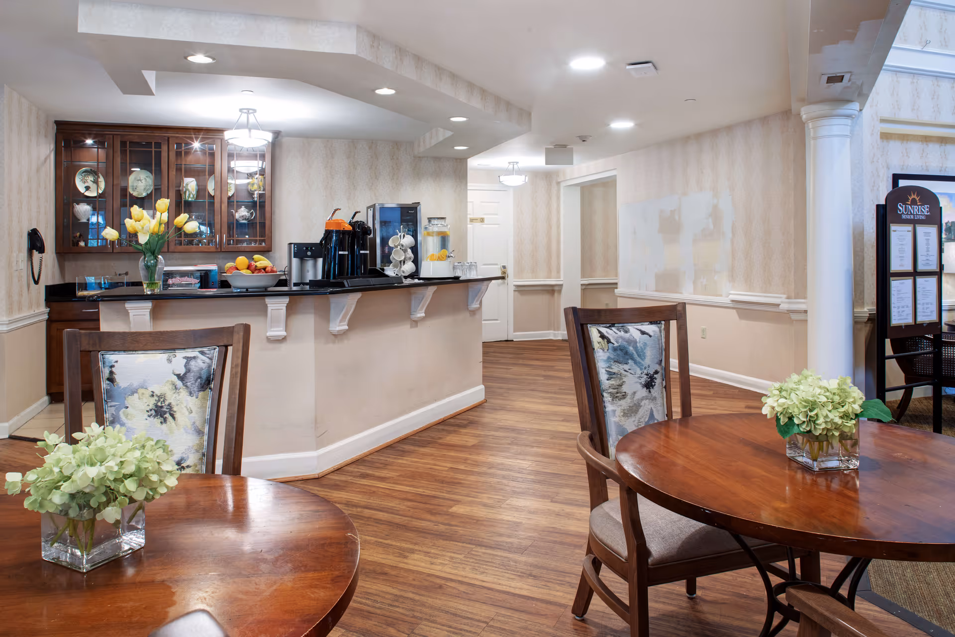 Cozy dining/lounge area with round wooden tables and chairs, a serving counter with a coffee station and flower arrangements, and wood flooring.