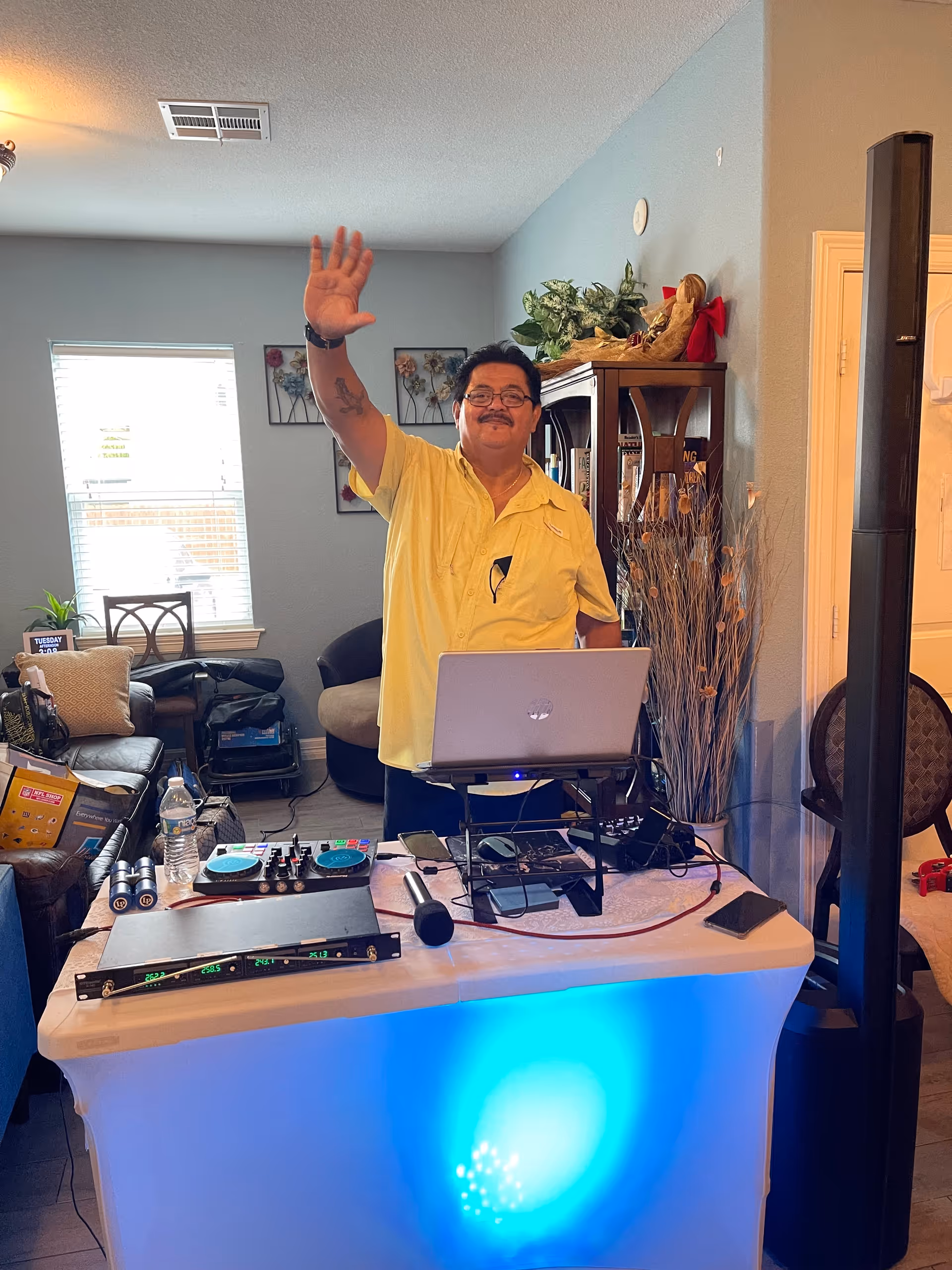 A man wearing a yellow shirt is standing behind a table with DJ equipment, including a laptop, microphone, and sound mixer, in a living room. He is smiling and waving with his right hand. The room has a couch, a window with blinds, wall art, and a wooden cabinet with plants and decorations on top.