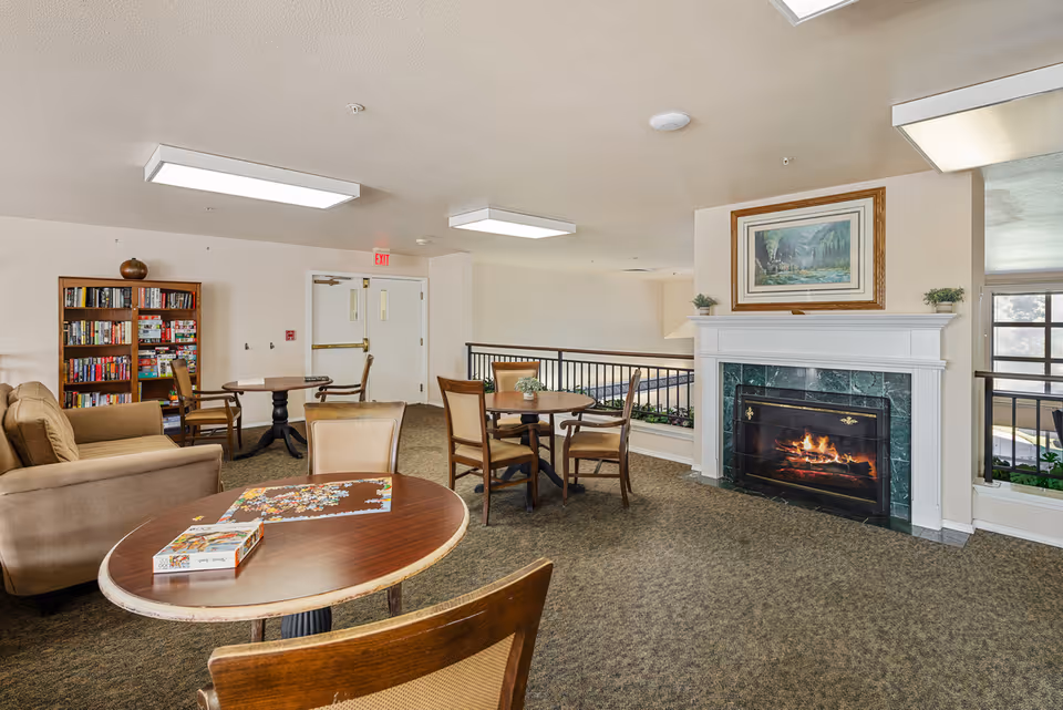 A cozy common area in a senior living facility featuring a lit fireplace with a framed painting above it, several round wooden tables with chairs, a puzzle in progress on one table, a bookshelf filled with books and board games, and a beige couch. The room has carpeted floors, white walls, and ceiling lights, with a railing overlooking a lower level.