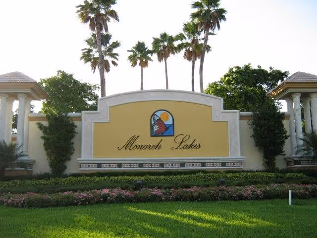 Entrance sign for Monarch Lakes featuring a decorative emblem with a sun and butterfly, surrounded by palm trees and well-maintained landscaping with green grass and flowering plants.