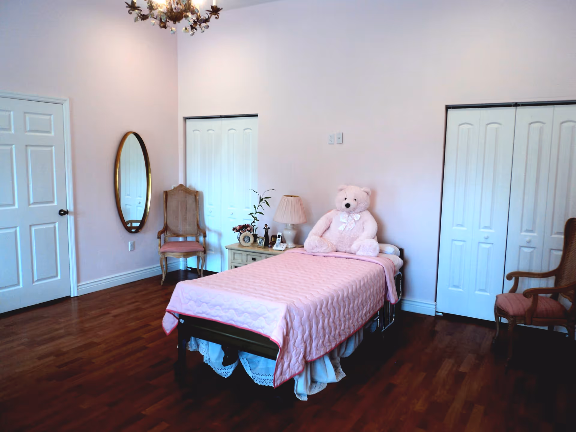 A bedroom with a single bed covered in a pink quilt and a large pink teddy bear sitting on it. The room has wooden flooring, light pink walls, two white closets with double doors, a wooden chair with a pink cushion, a small nightstand with a lamp, a phone, and decorative items. There is an oval mirror hanging on the wall and another wooden chair with a pink cushion on the right side.