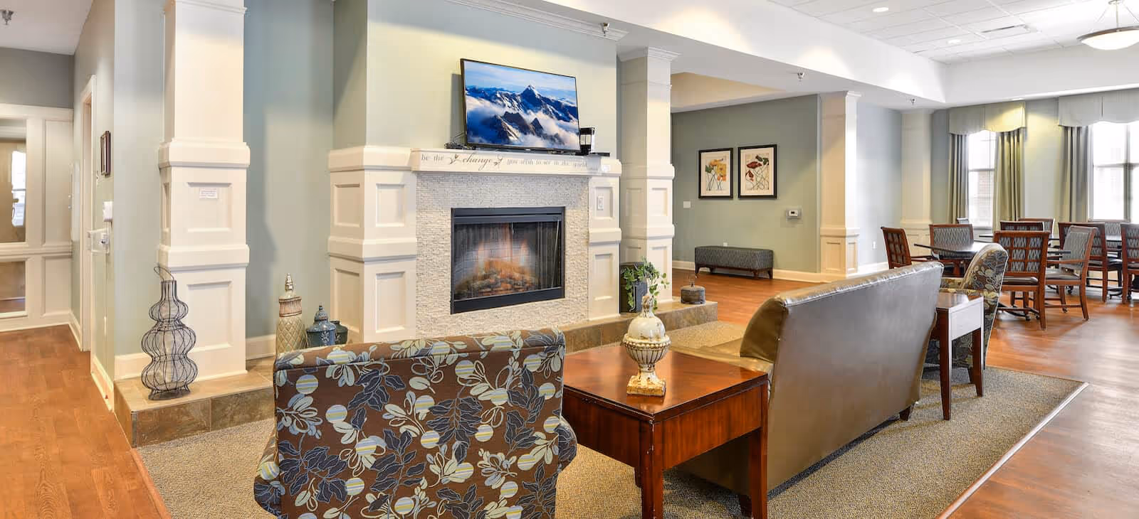 A cozy living room area in a senior living facility with a fireplace, a flat-screen TV mounted above it, and comfortable seating including a floral-patterned armchair and a leather sofa. There is a wooden coffee table with decorative items, and in the background, a dining area with tables and chairs near large windows letting in natural light.