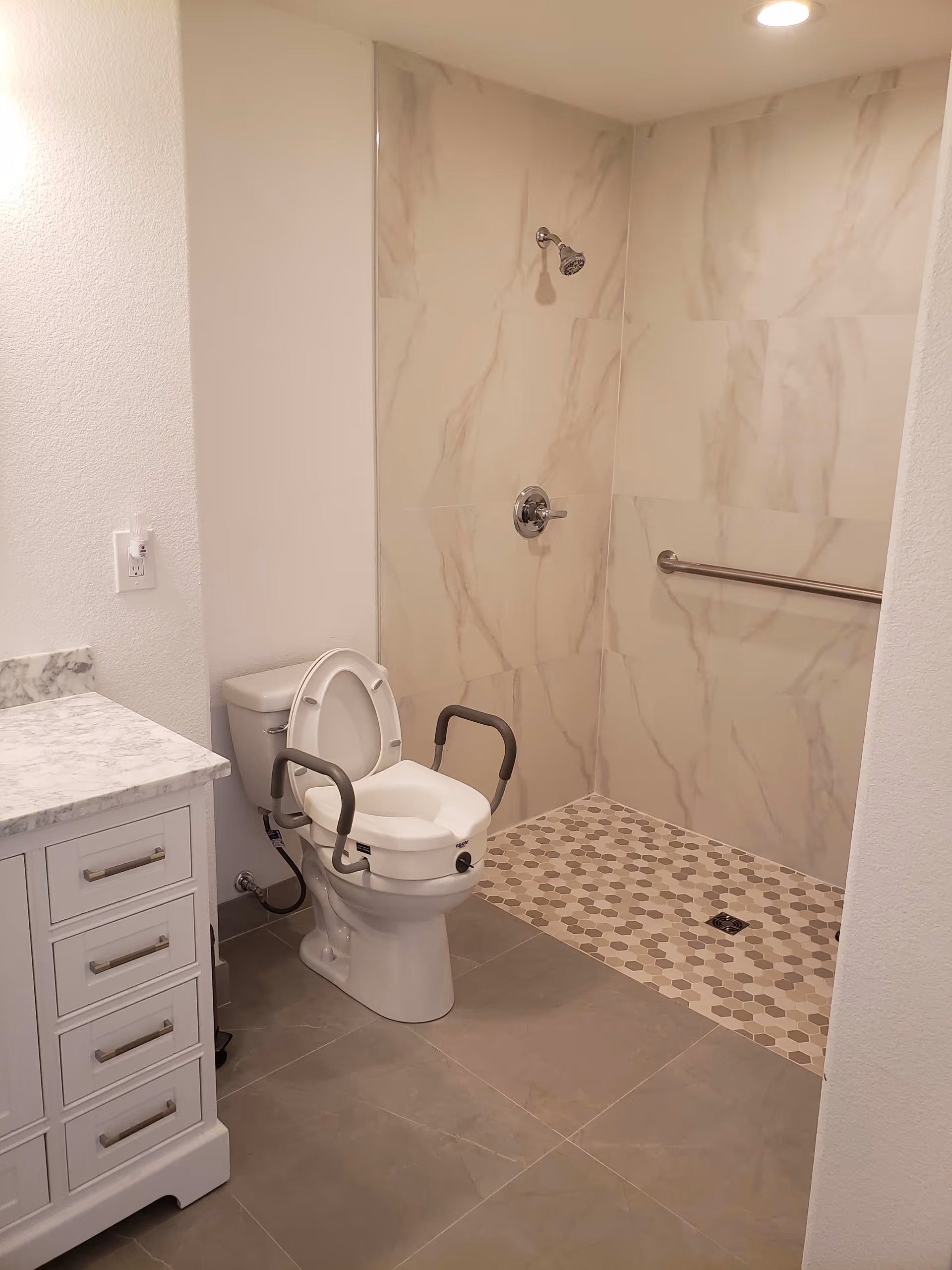 A bathroom with a white toilet equipped with safety rails and a raised seat. Next to the toilet is a white vanity with a marble countertop. The shower area has a wall-mounted showerhead, marble-patterned wall tiles, a grab bar, and a floor with hexagonal tiles in neutral tones. The floor outside the shower is tiled in large gray tiles.
