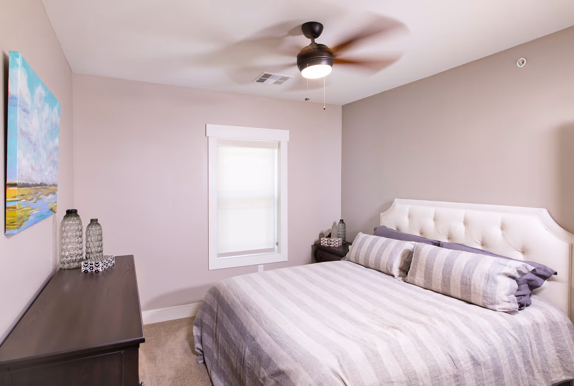 A bedroom with a large bed featuring a tufted white headboard and striped bedding in shades of gray and white. There is a dark wooden dresser with decorative glass bottles and a small box on top, a window with a white frame and a closed blind, a ceiling fan with a light, and a colorful landscape painting on the wall.