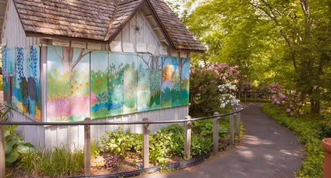A small wooden structure with a shingled roof and colorful painted panels depicting nature scenes, situated along a paved garden path surrounded by lush greenery and flowering bushes.