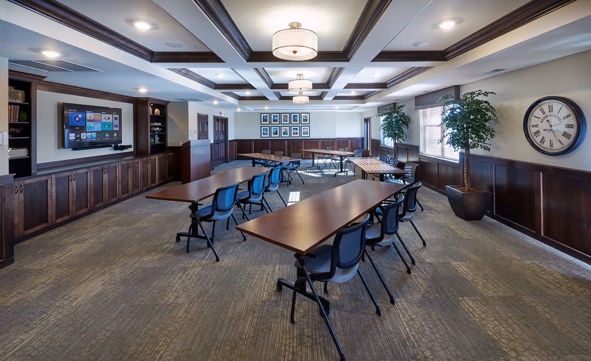 A well-lit meeting or activity room with multiple rectangular tables and blue chairs arranged in rows. The room features dark wood paneling and built-in cabinetry along one wall with a large flat-screen TV mounted above. There are large windows with blinds, two potted plants, a large wall clock, and framed pictures on the far wall. The ceiling has recessed lighting and decorative beams.
